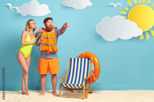 Pleased slim woman with bobbed hairstyle  wears bright green swimsuit  leans at shoulder of lover who has shocked expression  dressed in protective lifejacket  points into distance  pose at beach