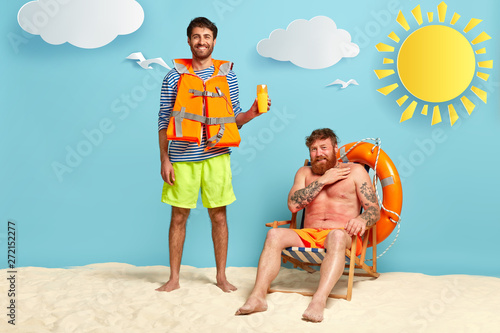 Photo of glad man suggests friend using sunscreen, has positive smile, wears lifejacket, upset red haired guy has sunburnt skin, sits at deckchair, needs sunblock lotion. Skincare and summer vacation