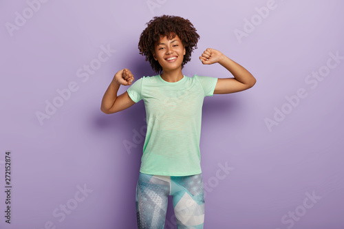 Half length shot of happy young Afro American woman stretches hands, does morning exercises with good mood, wears casual t shirt and leggings, has broad smile, isolated over purple background
