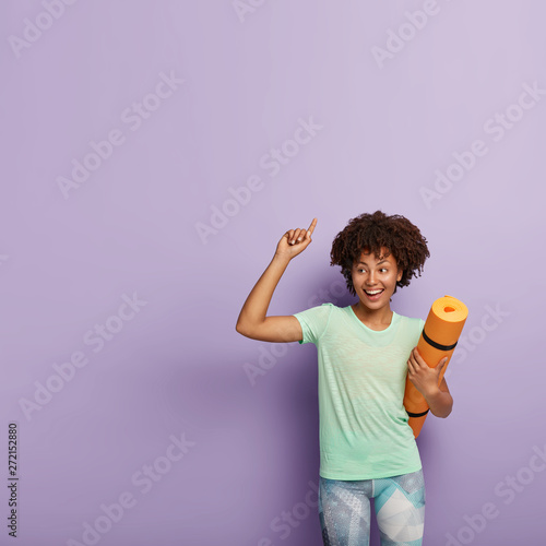 Positive dark skinned female with Afro hairstyle, wears casual t shirt and leggings, has fitness meditation, yoga training, points at blank space over purple background, shows direction to gym