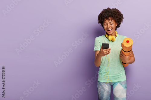 Joyous young girl has break in training, poses with fitness mat, holds smartphone, enjoys songs in headphones, wears sportswear, isolated over purple wall, copy space. Sportswoman listens music