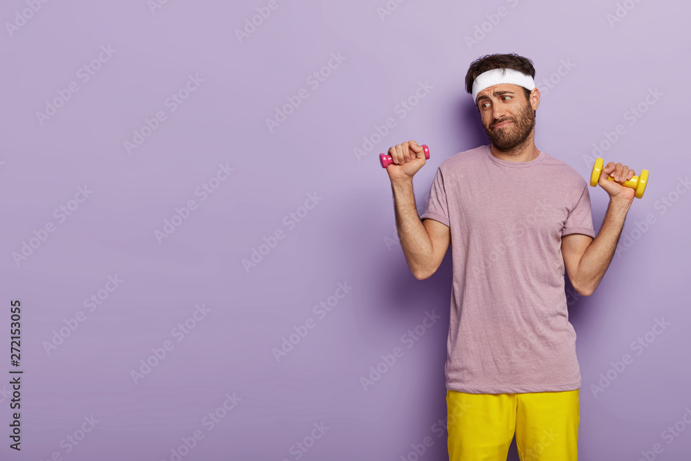 Young man looks unwillingly, holds dumbbells in both hands, has no desire for training, dresses in active wear, stands against purple background with copy space for your advertising content. Young man looks unwillingly, holds dumbbells in both hands, has no desire for training, dresses in active wear, stands against purple background with copy space for your advertising content.