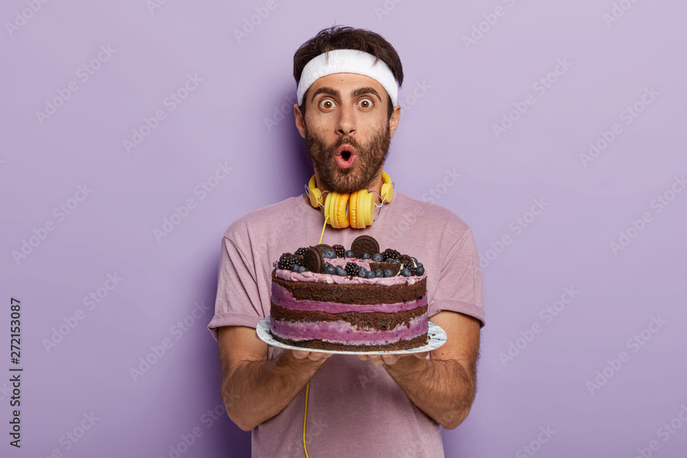 Astonished handsome guy with bristle, dark hair, stare at camera, has wide opened eyes and mouth, holds delicious alluring fruit cake, stereo yellow headphones on neck. Unhealthy eating concept Astonished handsome guy with bristle, dark hair, stare at camera, has wide opened eyes and mouth, holds delicious alluring fruit cake, stereo yellow headphones on neck. Unhealthy eating concept