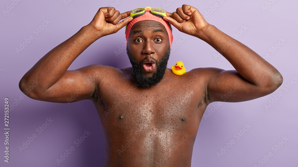 Photo of impressed swimming instructor keeps hand on goggles, shocked with bad weather conditions at beach, has dark skin, being wet. Surprised shirtless Afro American swimmer has active rest Photo of impressed swimming instructor keeps hand on goggles, shocked with bad weather conditions at beach, has dark skin, being wet. Surprised shirtless Afro American swimmer has active rest