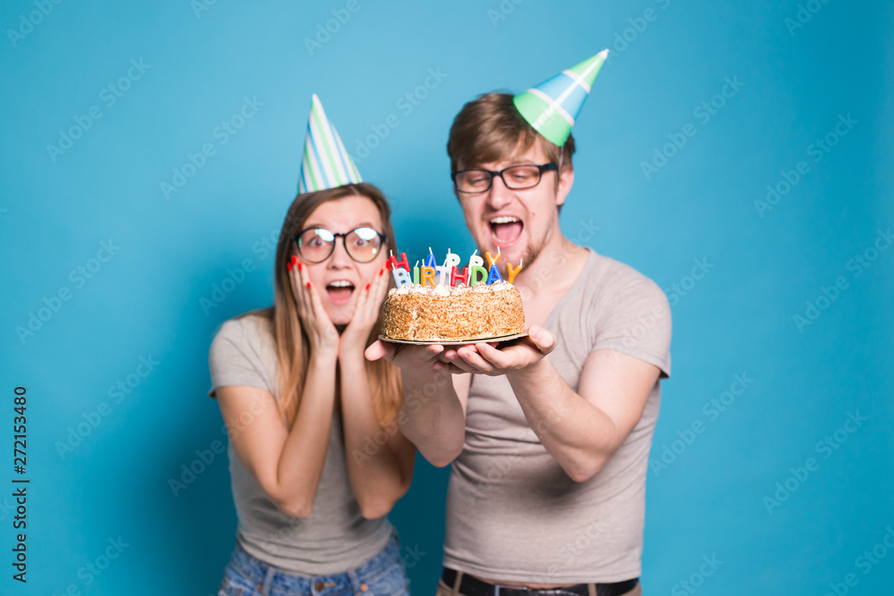 Funny young couple in paper caps and with a cake make a foolish face and wish happy birthday while standing against a blue background. Concept of congratulations and fooling around.