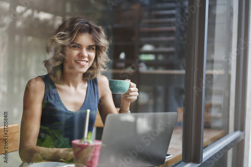 Successful lucky attractive european blond woman short curly hairstyle hold coffee cup working laptop edit photograph freelance, sit window bar cafe look outside search inspiration