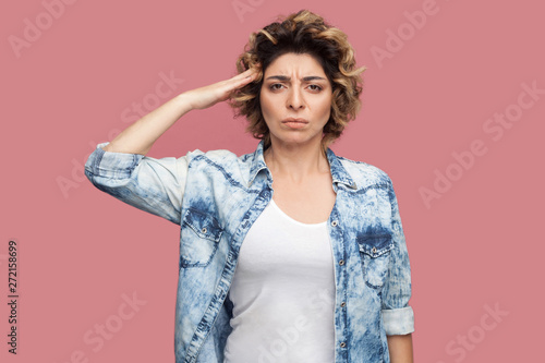 Yes sir. Portrait of serious young worker woman with curly hairstyle in casual blue shirt standing and looking at camera with salute and ready to do smoething. studio shot, isolated on pink background