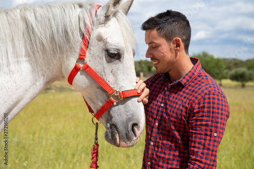 Side view of young male in checkered shirt gently touching head of white horse with red bridle while standing in countryside field on cloudy day