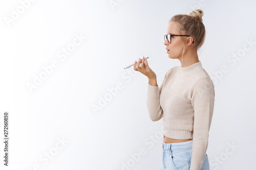 Profile shot of young stylish blond woman in glasses holding smartphone near mouth talking speaker recording voicemessage over white background with bossy serious-looking focused look photo