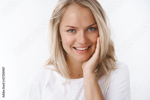 Close-up shot of satisfied pleased sensual blond woman touching soft and silk skin on cheek smiling delighted and sensual taking skincare vitamins and products washing off cosmetics over white wall