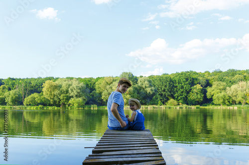 Father and a tiny girl in dungarees are sitting on the bridge and looking at the camera. Back view, copy space.