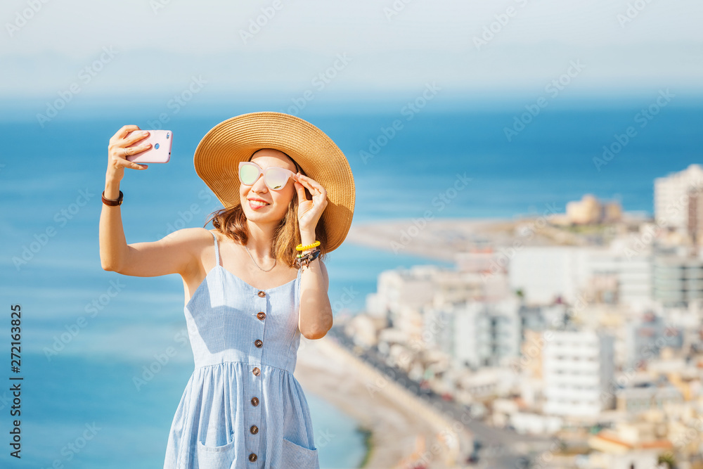 A young stylish girl in a big hat taking selfie near the sea resort town, aerial view