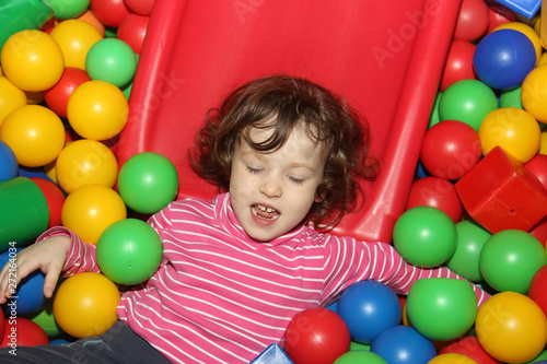 Little curly cute smile girl plays in balls for a dry pool. Play room. Happiness.