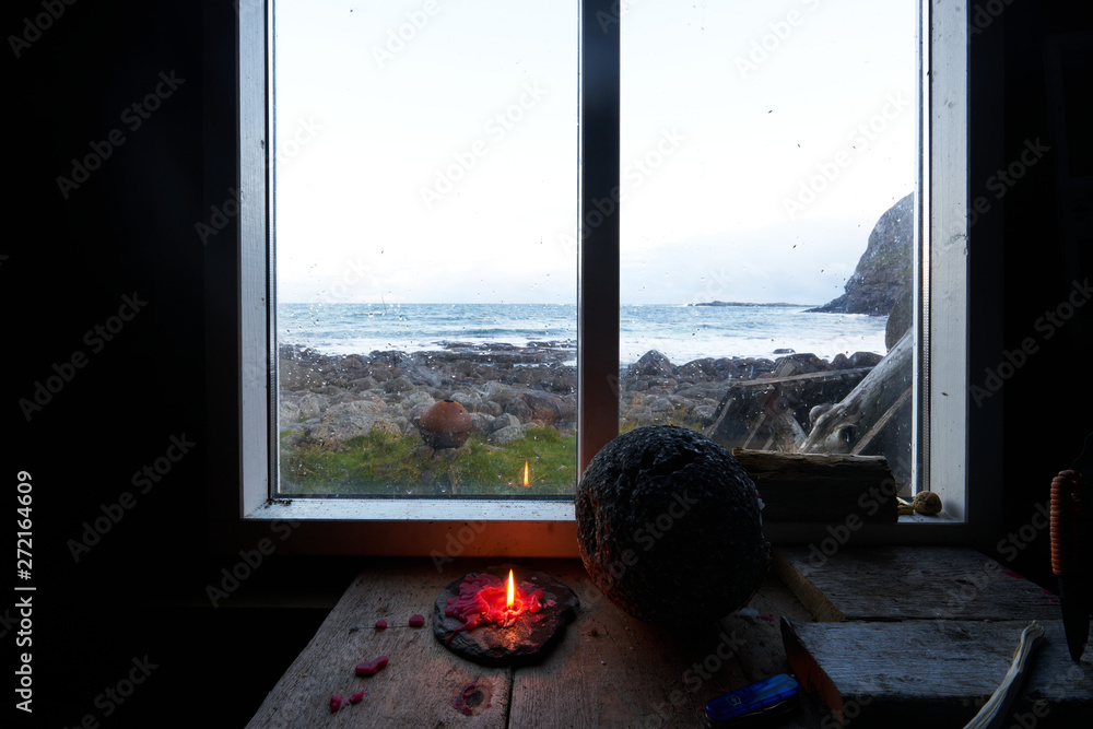 Look through the window of a cabin at the coast with a burning candle in the foreground.