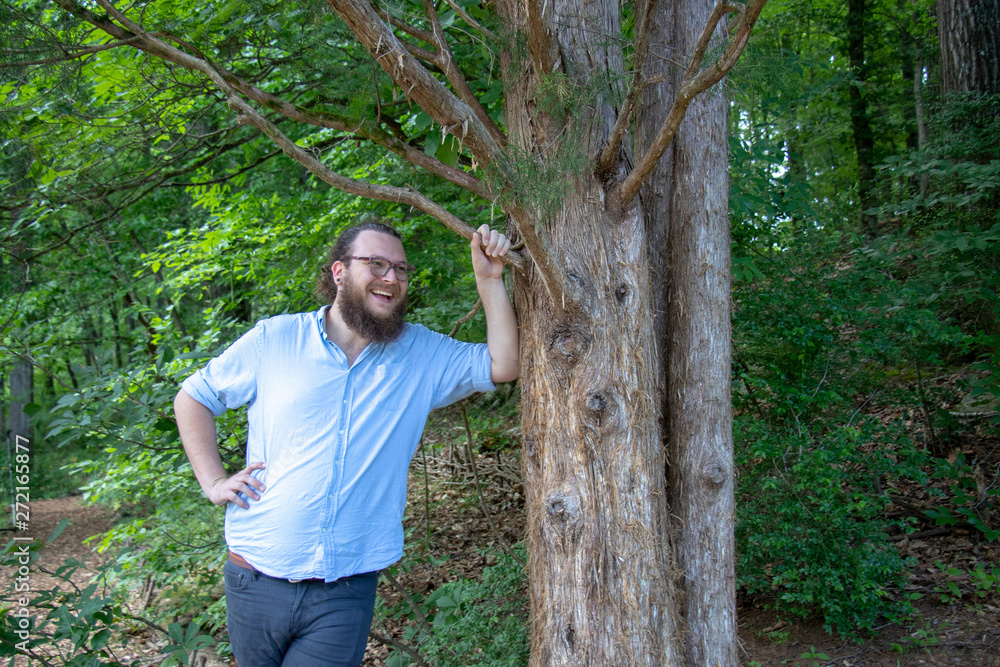 Smiling young bearded man leans against a tree. Relaxed hipster with beard, earring and ponytail in a natural environment. Smiling young bearded man leans against a tree. Relaxed hipster with beard, earring and ponytail in a natural environment.