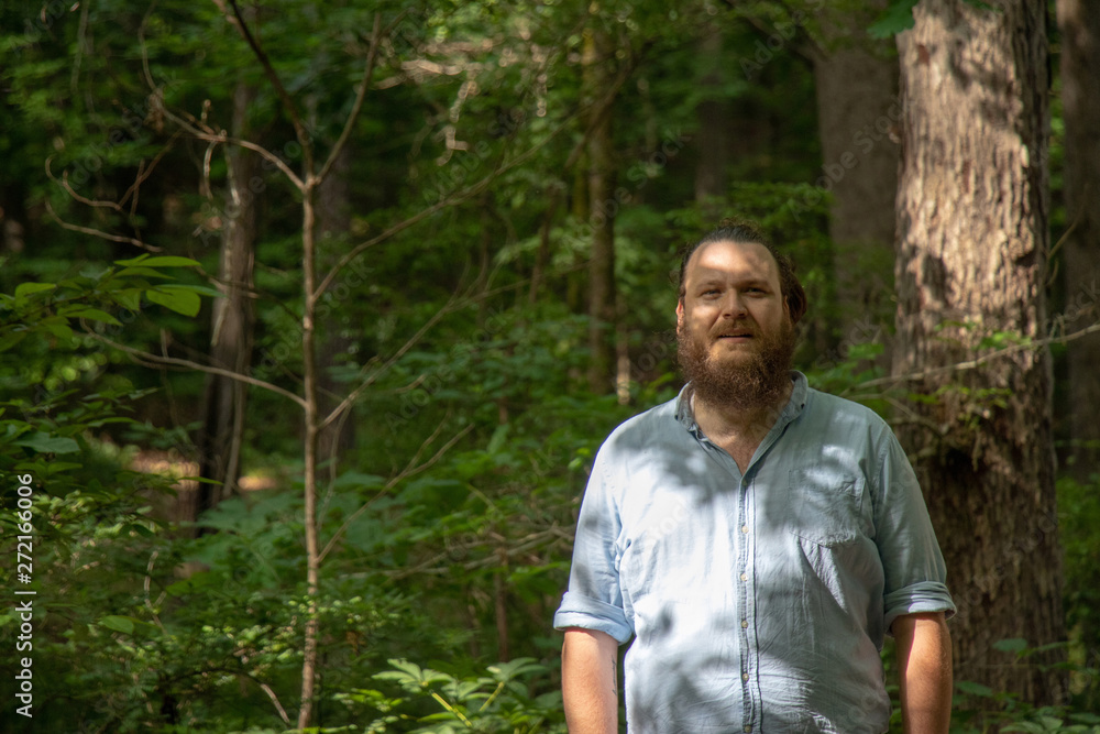Young bearded man standing in the woods in dappled shade looks thoughtfully at the camera. Relaxed hipster in a natural environment. Young bearded man standing in the woods in dappled shade looks thoughtfully at the camera. Relaxed hipster in a natural environment.