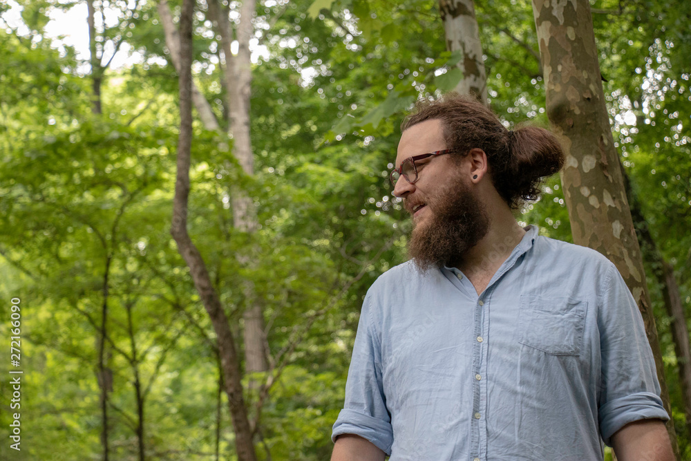 Young man looking around in forest is about to smile. Relaxed hipster with glasses, beard and ponytail in a natural environment. Young man looking around in forest is about to smile. Relaxed hipster with glasses, beard and ponytail in a natural environment.