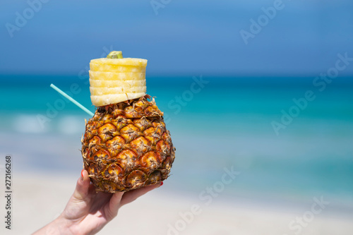 Woman hand holds tropical pineapple cocktail on the beach. Beautiful sea ocean sky white sand beach. Cuba space for text