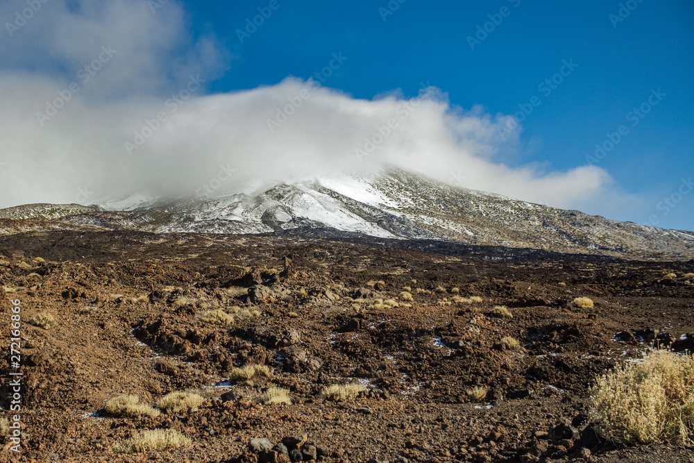 Mountain Teide with white snow spots, partly covered by the clouds. Bright blue sky. Teide National Park, Tenerife, Canary Islands, Spain.