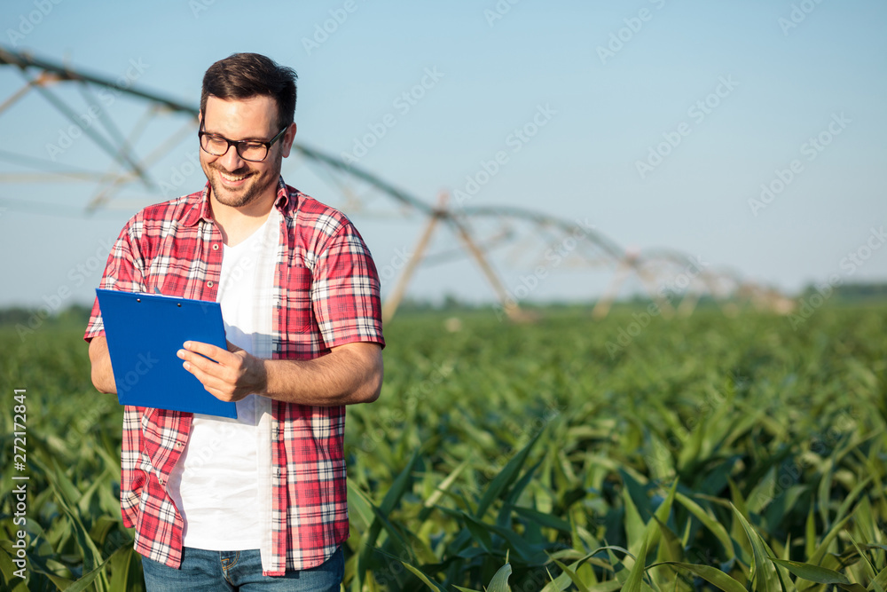 Happy young farmer or agronomist in red checkered shirt writing on a clipboard, inspecting a corn field. Irrigation system in the background. Organic farming and food production