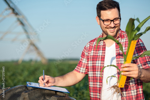 Happy young farmer or agronomist in red checkered shirt measuring young corn plant stem size with a ruler, writing data to a questionnaire. Organic farming and healthy food production.