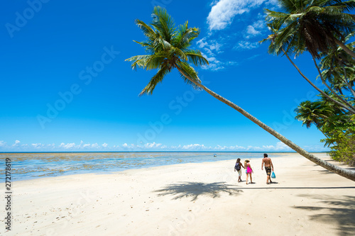 Figures walking under a palm tree leaning out over a wide empty beach on a remote tropical island in Bahia, Brazil