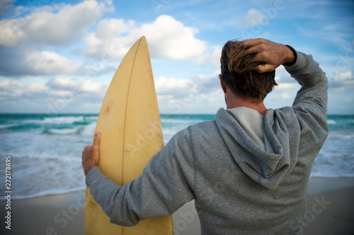Surfer in gray hoodie holding his surfboard on the shore of a windy tropical beach