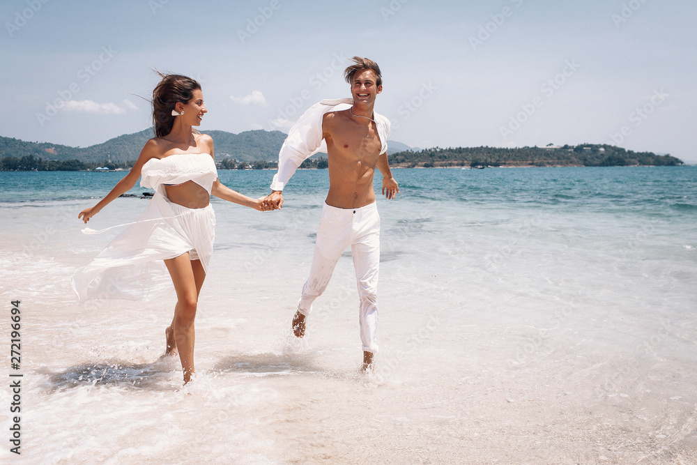 Young beautiful man and woman couple in white clothes running along the beach. Couple man and woman in the rays of the sun on the ocean bank.