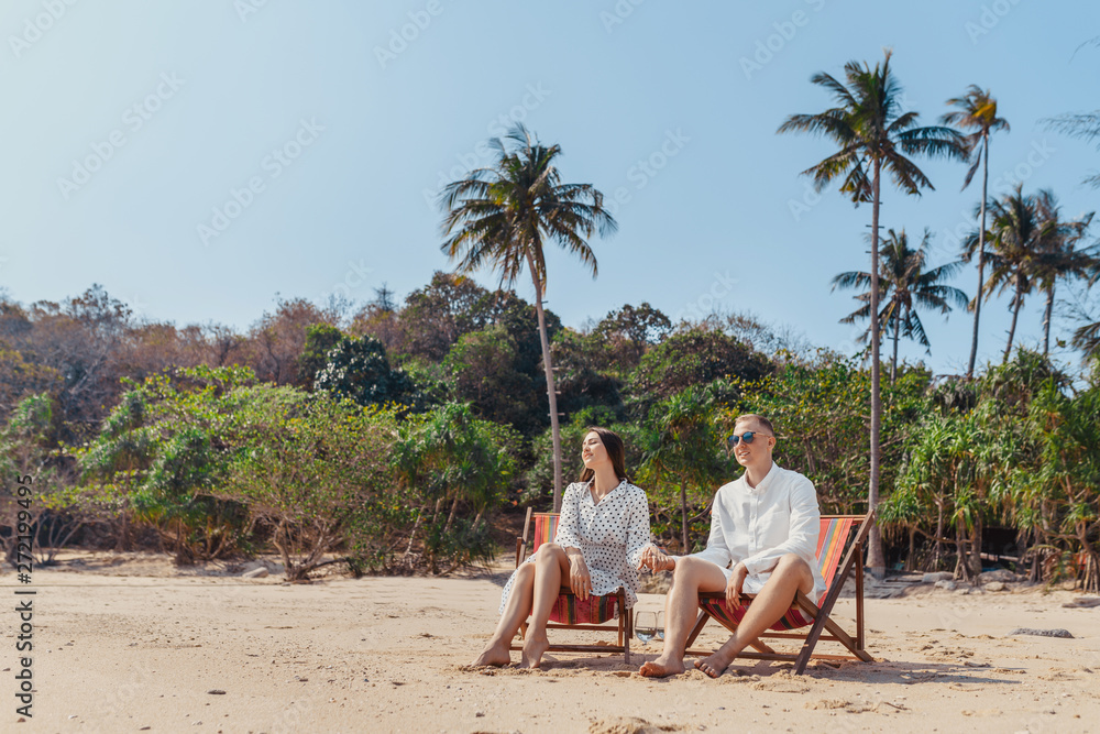 Smiling partners hold hands while resting on comfortable chairs by sea beach. Palm trees on the background