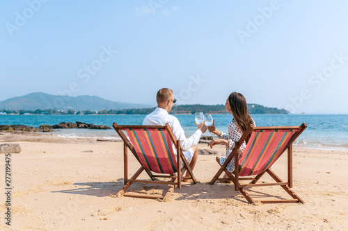 Smiling partners enjoying drinks champagne while resting on comfortable chairs by sea beach. Blue sea on the background. Thailand. Back view