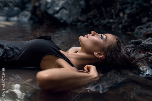 Close up photo of an Attractive woman with ideal beautiful face enjoying nature lying on her back in darkness lost in thought thinking deeply amongst dark forest and black stones #272201819