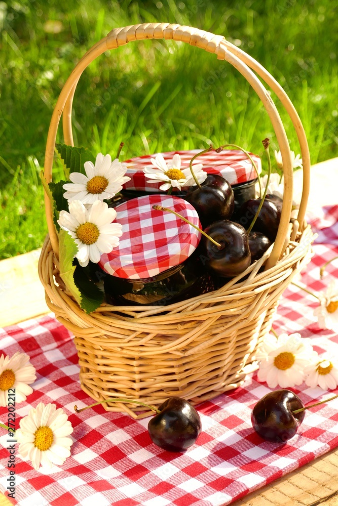 Cherry jam. sweet cherry jam in glass jars in a wicker basket, cherry and chamomile on a red checkered napkin on a wooden table on a green background.Homemade jam. Canned berries. Cherry jam. sweet cherry jam in glass jars in a wicker basket, cherry and chamomile on a red checkered napkin on a wooden table on a green background.Homemade jam. Canned berries.