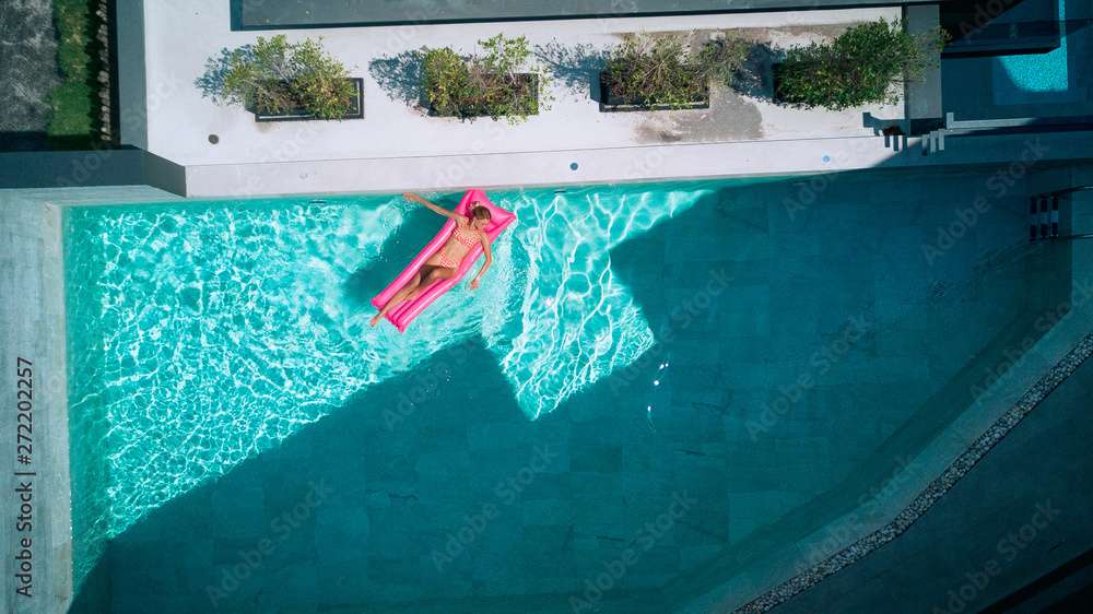 Beautiful young lady relaxing in swimming pool, swims on inflatable mattress and has fun in water on  vacation, tropical holiday resort, aerial drone view from above