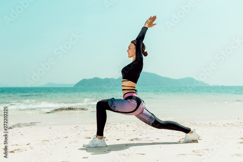 Beautiful woman practices yoga by the sea on a sunny day. The woman does stretching exercises. Dumbbells lying on the sand.