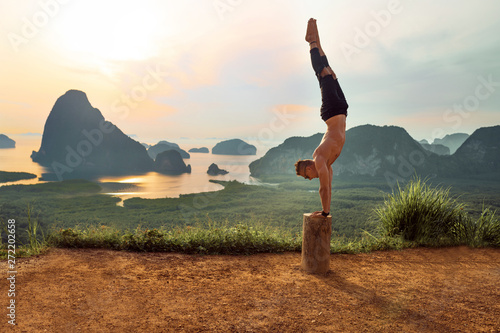 Happy man in black clothes doing yoga pose standing on his hands on the tree. Incredible sunset background with mountains and lakes. The concept of balance and harmony in life.  photo