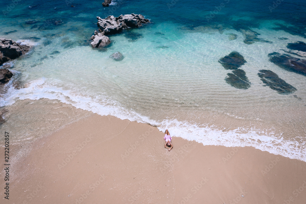 Obraz premium Young woman in a dress lying on the back on the white sand near the waves of blue sea. Top view, Andaman Sea, Phuket, Thailand. Aerial