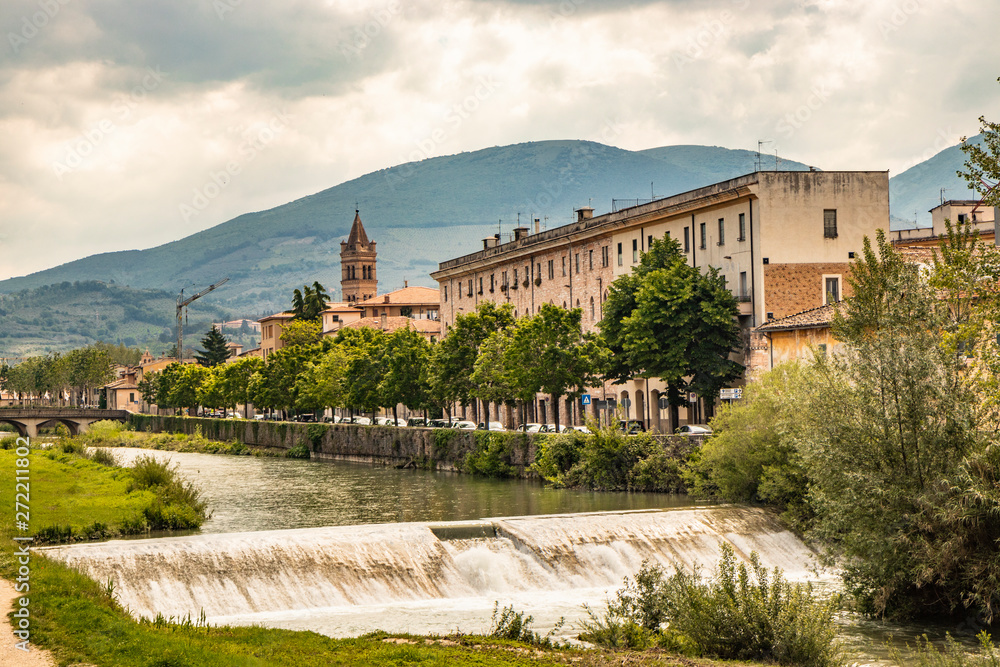 A view of Foligno, crossed by the river Topino, a bell tower rises above the roofs of the houses. The cloudy sky at sunset. The canal, the bridge and the trees.