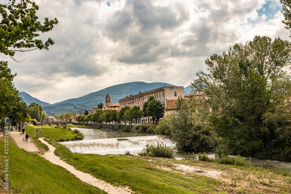 A view of Foligno, crossed by the river Topino, a bell tower rises above the roofs of the houses. The cloudy sky at sunset. The canal, the bridge, the trees. Some people walk in the park with the dog.