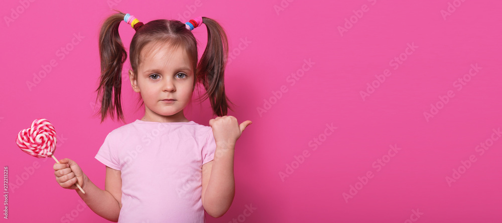 Close up portrait of little cute child, looking directly at camera, holding heart bright lollipop, pointing aside with her thumb, charming kid wearing casual rose t shirt, posing isolated on rose.