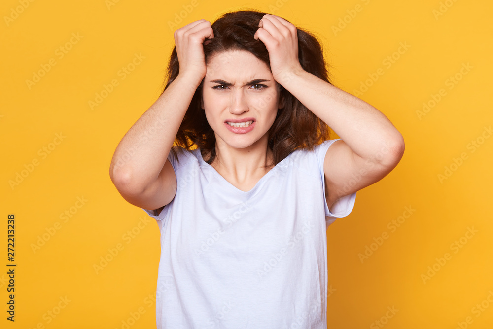 Indoor studio shot of stressed exhausted woman having unpleasant facial expression, touching her head with hands, pulling her hair out, posing with irritation in her eyes. Stressful work concept. Indoor studio shot of stressed exhausted woman having unpleasant facial expression, touching her head with hands, pulling her hair out, posing with irritation in her eyes. Stressful work concept.