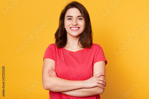 Indoor shot of magnetic beautiful young girl standing with folded arms, looking directly at camera, having sincere smile on her face, posing straight isolated over yellow background in studio.