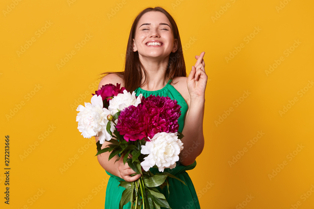 Stidio shot of pretty Caucasion woman raises crossed fingers, prays for good luck, has pleased and happy expression, wears green sundressr, holds white and burgundy peonies, believes in fortune. Stidio shot of pretty Caucasion woman raises crossed fingers, prays for good luck, has pleased and happy expression, wears green sundressr, holds white and burgundy peonies, believes in fortune.