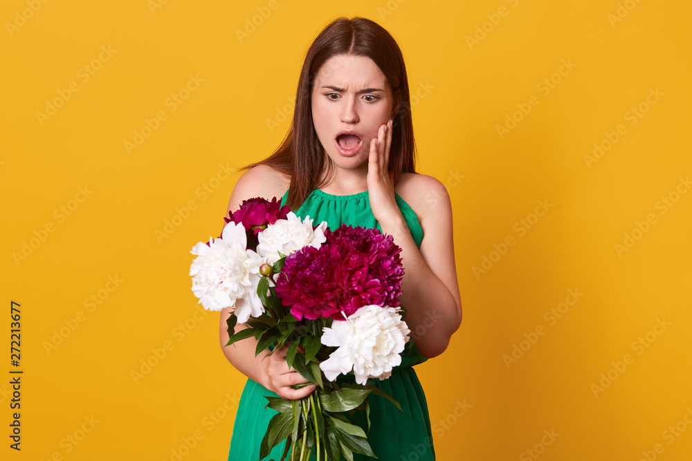 Beautiful girl standing with bouquet of burgundy and white peonies in her hands, wearing green sundress, posing with open mouth, has astonished facial expression, isolated over yellow background. Beautiful girl standing with bouquet of burgundy and white peonies in her hands, wearing green sundress, posing with open mouth, has astonished facial expression, isolated over yellow background.