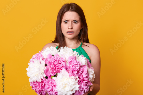 Worried shocked young woman looking at pink and white flowers with surprise, holding peonies in both hands, opening eyes widely, having unpleasant facial expression, wearing green summer dress.