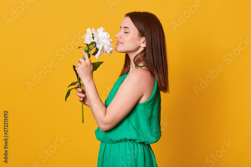 Close up profile portrait of lady wearing green sundress keeps beautiful flowers in hands on yellow studio background, being happy to recive peonies as gift. Copy space for advertisment or promotin. photo