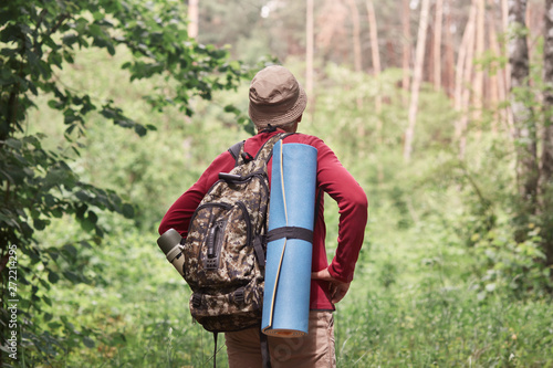 Back view of active backpacker looking for way out in forest, having sleeping pad and dark rucksack with thermos at his back, enjoying active holidays, stick to travel lifestyle. Travel concept.