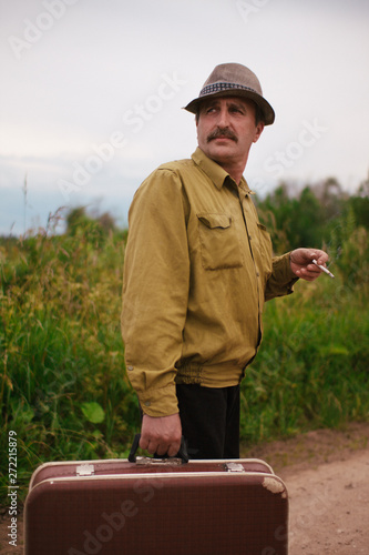 An elderly man travels with a suitcase. A villager is on the road to a long way and smoking a cigarette.
