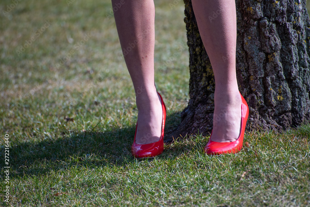 Legs with a woman's red shoes on the grass, a tree in the side. Concept of feminine sensuality.