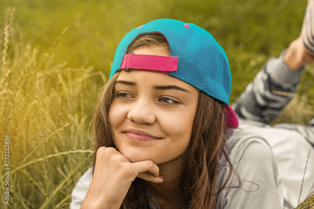 A beautiful teen girl in a baseball cap with her hair down lying on the grass looks away and smiles. A beautiful teen girl in a baseball cap with her hair down lying on the grass looks away and smiles.