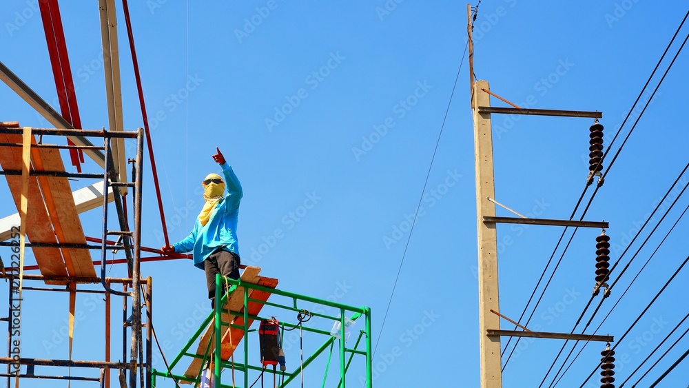 Low angle view of construction worker in sunglasses and yellow veil on high scaffold is working to build up roof building structure with electric pole and blue clear sky background, risky work cocept Low angle view of construction worker in sunglasses and yellow veil on high scaffold is working to build up roof building structure with electric pole and blue clear sky background, risky work cocept